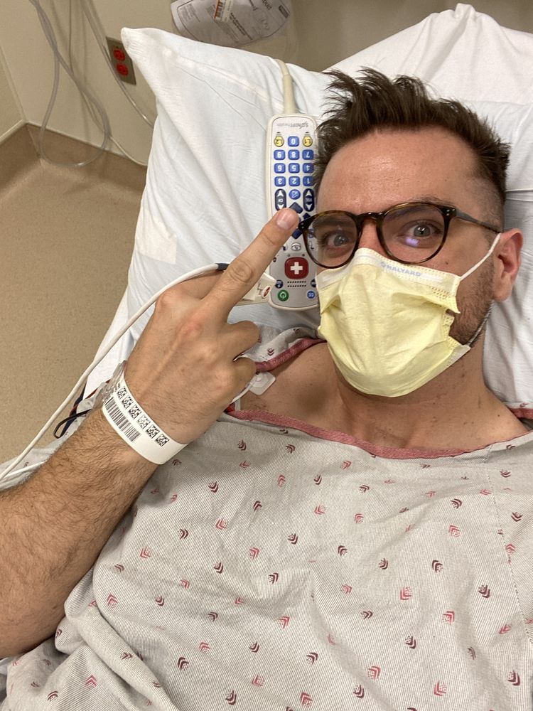 Rob in a hospital bed wearing a mask, giving a thumbs up
